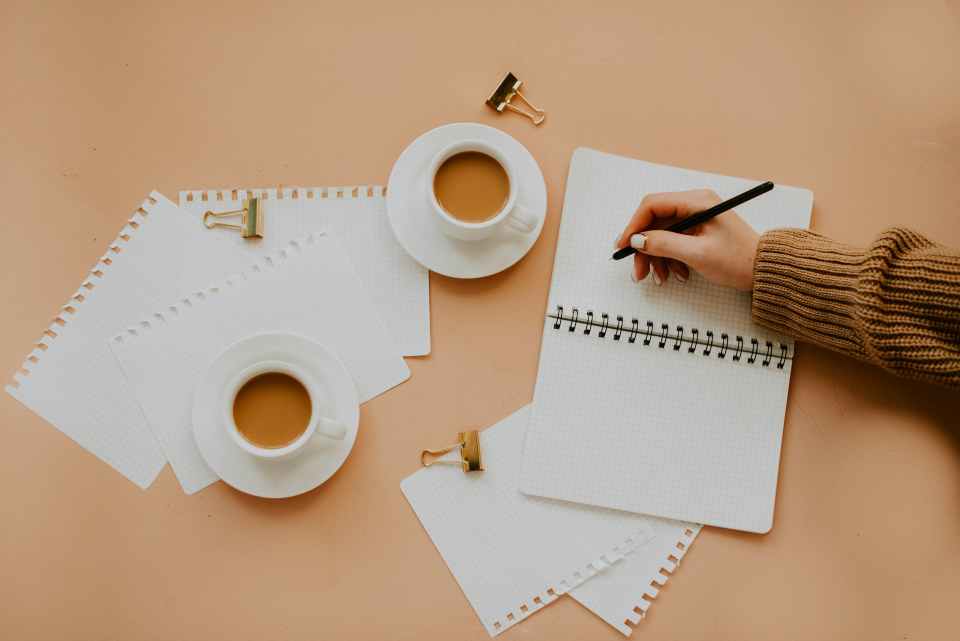 Woman writing in a notebook with papers spread around and two cups of coffee.