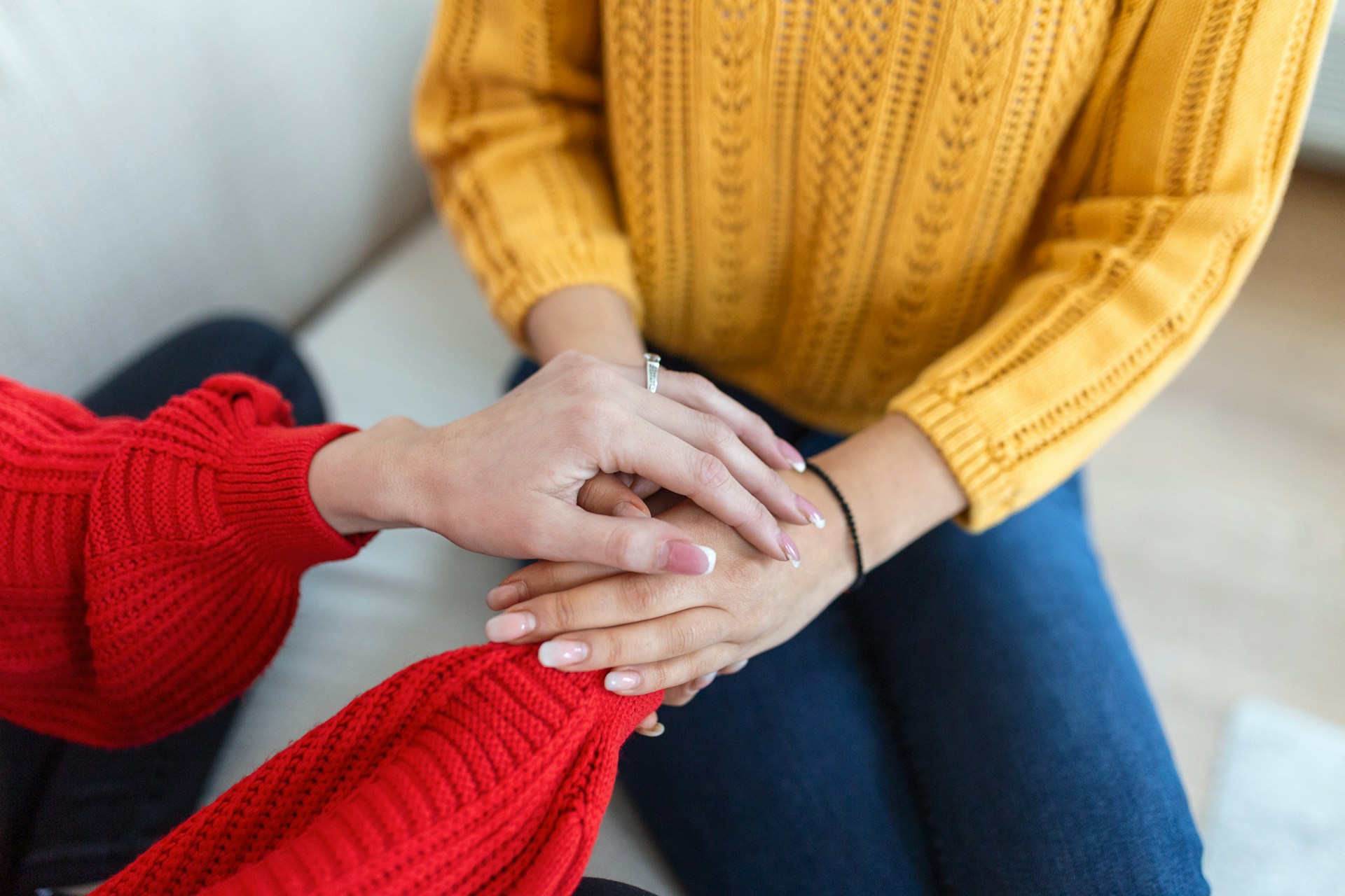 Two women holding hands to give comfort, only hands and torsos are shown one has a yellow sweater and the other a red sweater