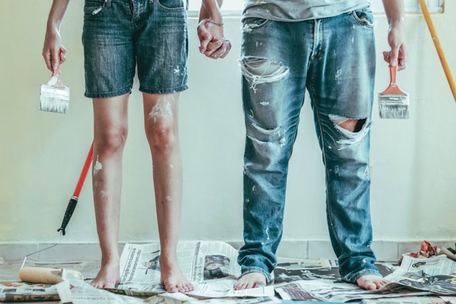 Man and Woman from the waist down wearing jeans and covered in paint holding paint brushes just finishing a DIY project.